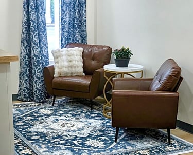 Two brown leather armchairs on a blue patterned rug with a white side table and damask curtains.
