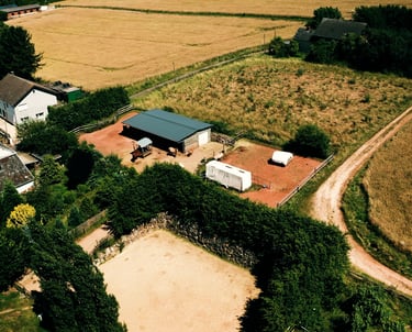 Luftaufnahme eines ländlichen Hofes mit Stall, umzäuntem Reitplatz und angrenzenden Feldern