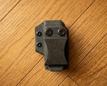 Black Kydex IWB magazine carrier with belt clip on a light oak wood background.