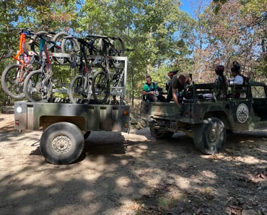 Military vehicle towing a bike rack trailer loaded with mountain bikes for riders on a wooded trail.