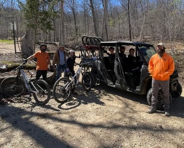 A group of mountain bikers with their gear and a black shuttle utility vehicle on a wooded trail.