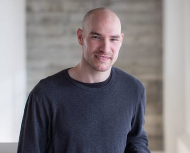 Professional bald man in a casual grey sweater posing for a portrait in a modern office.