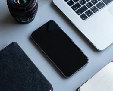Clean, flat-lay photography of a smartphone, a sleek laptop, and a notepad on a light gray desk. Muted blue tones and sharp focus.