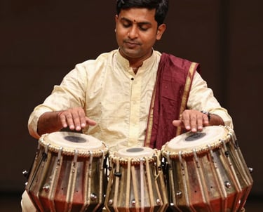 A professional South Asian / Indian male musician playing a set of traditional tabla drums, focused expression, wearing ethnic attire in Soft Cream and Deep Burgundy colors.