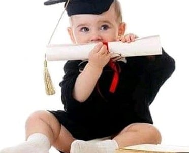 A baby wearing a graduation cap and gown holding a diploma and sitting next to a book.