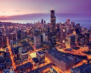 Aerial view of the Chicago IL skyline at sunset with illuminated skyscrapers and city lights.