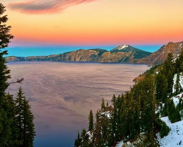 Sunset over Crater Lake National Park with snow-covered slopes and a pink sky over the deep blue caldera.