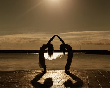 Silhouette of two people practicing yoga together on a coastal pier during a golden sunset.