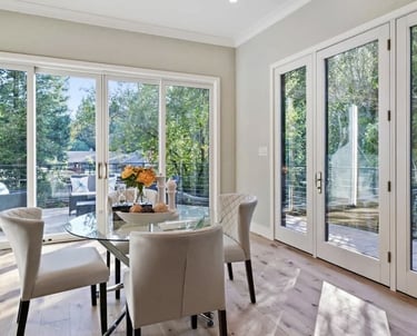 Dining area with a glass table and large windows overlooking an outdoor space.