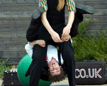 Two street performers in colorful costumes demonstrate a vertical acrobatic juggling balancing act. 
