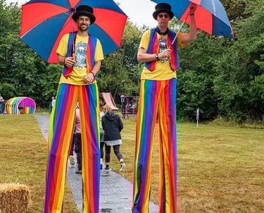 Two circus entertainers on stilts wearing rainbow trousers and top hats with umbrellas at an outdoor festival.