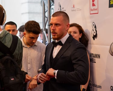 A man in a navy suit and bowtie poses on a red carpet in front of a brand sponsor backdrop.