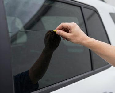 A skilled technician carefully applying window tint film to a sleek black car under bright workshop lights.