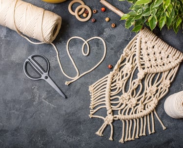 rollo de hilo, tijeras, cuentas y telar de macrame sobre una mesa con una planta