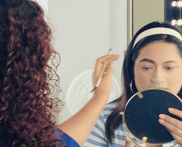Maquilladora profesional aplica lápiz de cejas a una mujer que sostiene un espejo en un estudio.