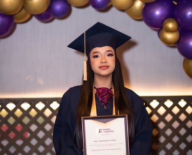 Una graduada sonriente con toga y birrete azules sostiene su diploma bajo un arco de globos morados 