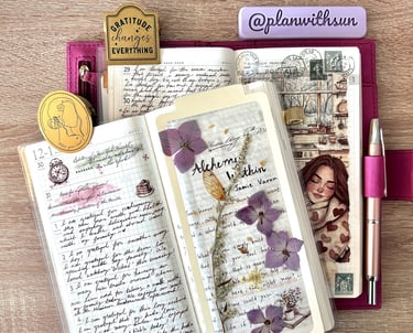 Open journaling notebook with handwritten entries, dried purple flowers, and stationery on a desk.