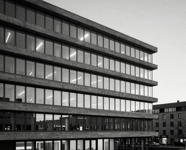 A black and white photography of a minimalist concrete office building during twilight. Sharp horizontal lines, glowing windows. Western European city.