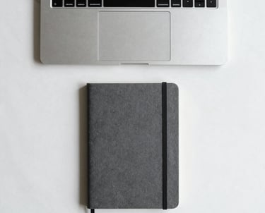 Top-down shot of a minimalist workspace with a single brushed silver laptop and a charcoal grey notebook on a white marble desk. Perfectly aligned, zero clutter. International aesthetic.
