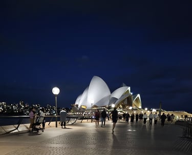 Night view of the Sydney Opera House illuminated with crowds walking along the harbor boardwalk.