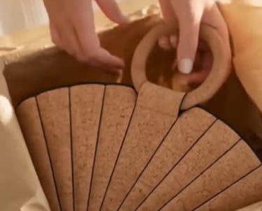 A person unboxing a fan-shaped cork handbag with round wooden handles from brown tissue paper.