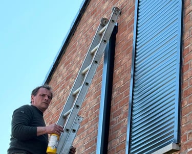 A technician on a ladder installs modern black security roller shutters on a brick residential house.