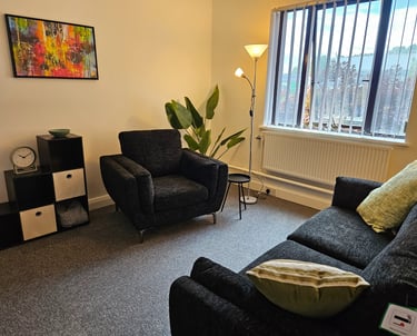 Modern cozy therapy room featuring dark fabric armchairs, colorful wall art, and indoor plants by a window.