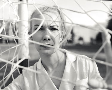 Black and white portrait of a woman looking through a soccer goal net on a field.