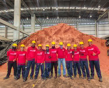 Industrial warehouse workers in red uniforms and yellow hard hats standing before a large pile of mineral ore.