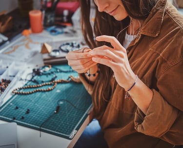 mujer sosteniendo joyeria artesanal frente a mesa de trabajo llena de cuentas e hilo