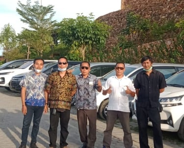 A group of five men standing in front of parked cars outdoors wearing batik shirts and sunglasses.