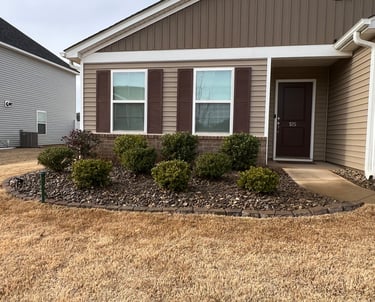 Flower bed in front of brown house with evergreen shrubs, cane rock for mulch and brown stone border.