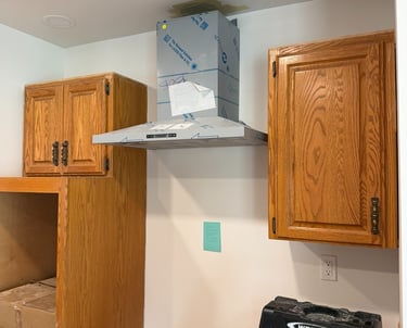 A stainless steel range hood being installed between oak kitchen cabinets during a home remodel.