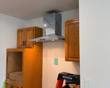 A stainless steel range hood installation in a kitchen remodel with oak cabinets and a Werner stepladder.