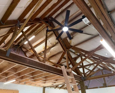 Exposed wood roof trusses in a garage attic with a black ceiling fan and LED shop lights.