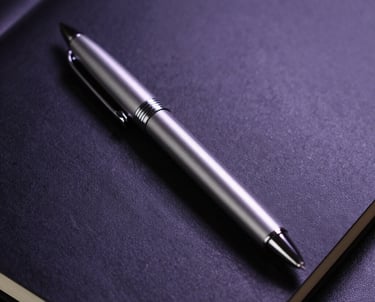 A close-up of a silver designer pen resting on a matte black notebook. The lighting is cool, with a single purple spotlight highlighting the texture of the paper.