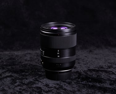 A minimalist studio portrait of a professional-grade camera lens on a black velvet surface. Soft purple light reflects on the glass. The composition is clean and centered.