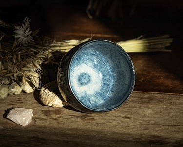 A handmade blue glazed ceramic tea bowl on a rustic wooden table with dried botanicals.