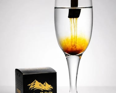 Golden Shilajit resin dissolving in a glass of water next to its black and gold branded packaging.