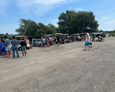 A group of people and families gathering by a row of parked golf carts in a gravel parking lot outdoors.