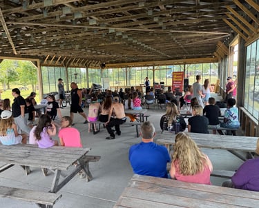 Families gather at a campground pavilion for a live magic show performance on a sunny afternoon.