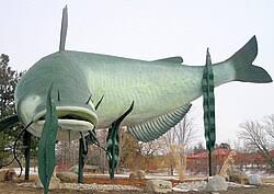 A giant channel catfish statue sculpture outdoors at a park with trees and rocks.