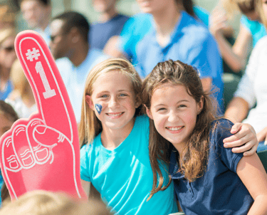 young girls in the stands of athletic event cheering on their team with a foam finger and big smiles