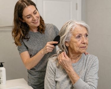 An elderly person sits at a dining table, holding a cup. The table is set with various dishes, pastries, and cups, indicating a meal. The environment appears cozy with natural light coming in through a window, and a houseplant is visible in the background.
