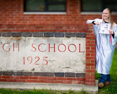 A blonde high school graduate in a white cap and gown posing by a brick school sign.
