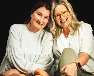 Two smiling women sitting closely together in white shirts against a dark background.