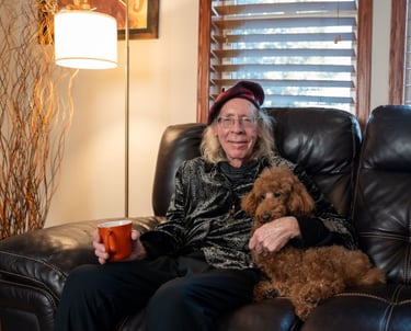 A senior man wearing a beret sitting on a leather sofa with a brown toy poodle and a coffee mug.