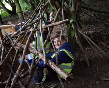 Cub Scout sitting inside a den made from sticks during a forest survival activity.