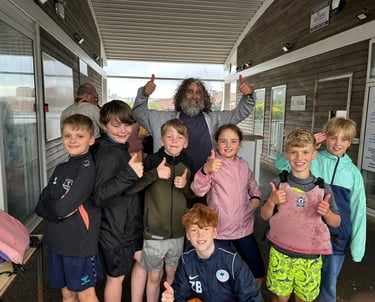 Group of Cub Scouts giving thumbs up with a scout leader under a shelter at a waterfront venue.