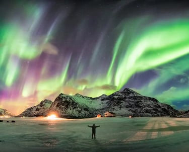 A person stands under the vibrant green and purple aurora borealis over snowy mountains in Norway.